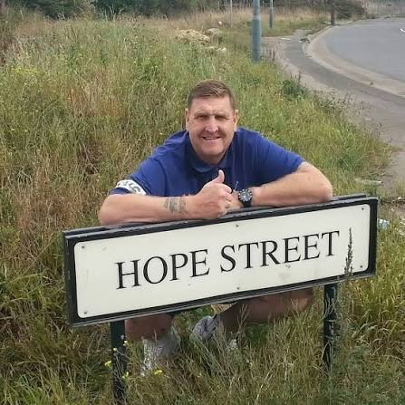 Gram Seed smiling and giving a thumbs up behind a Hope Street road sign